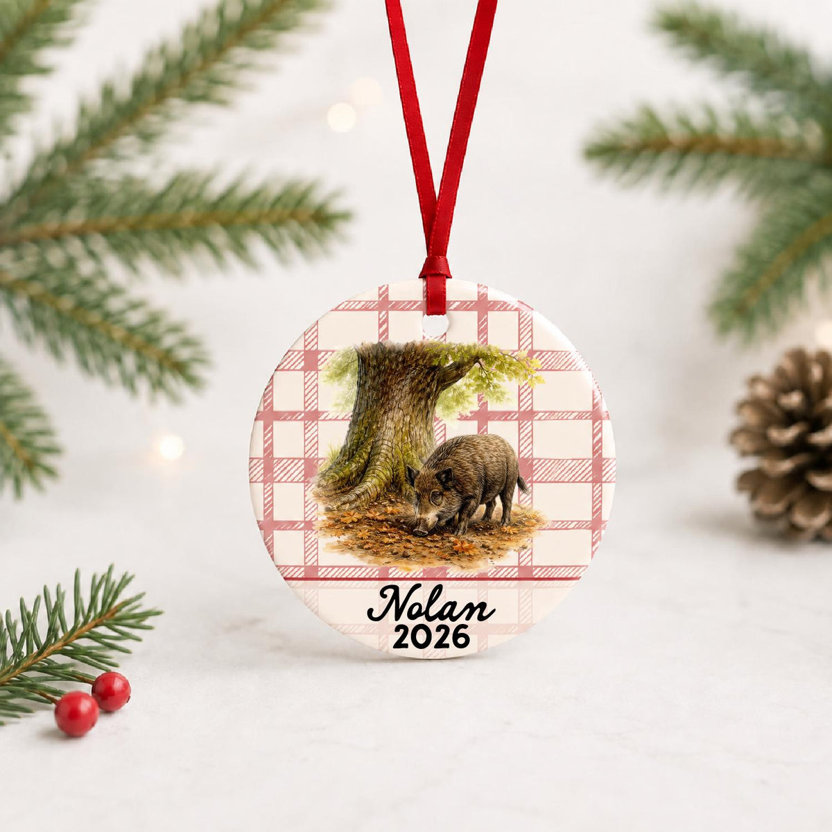 Personalized wild boar ceramic ornament with name and year on red plaid with red ribbon on a white surface with pine branches and pinecones