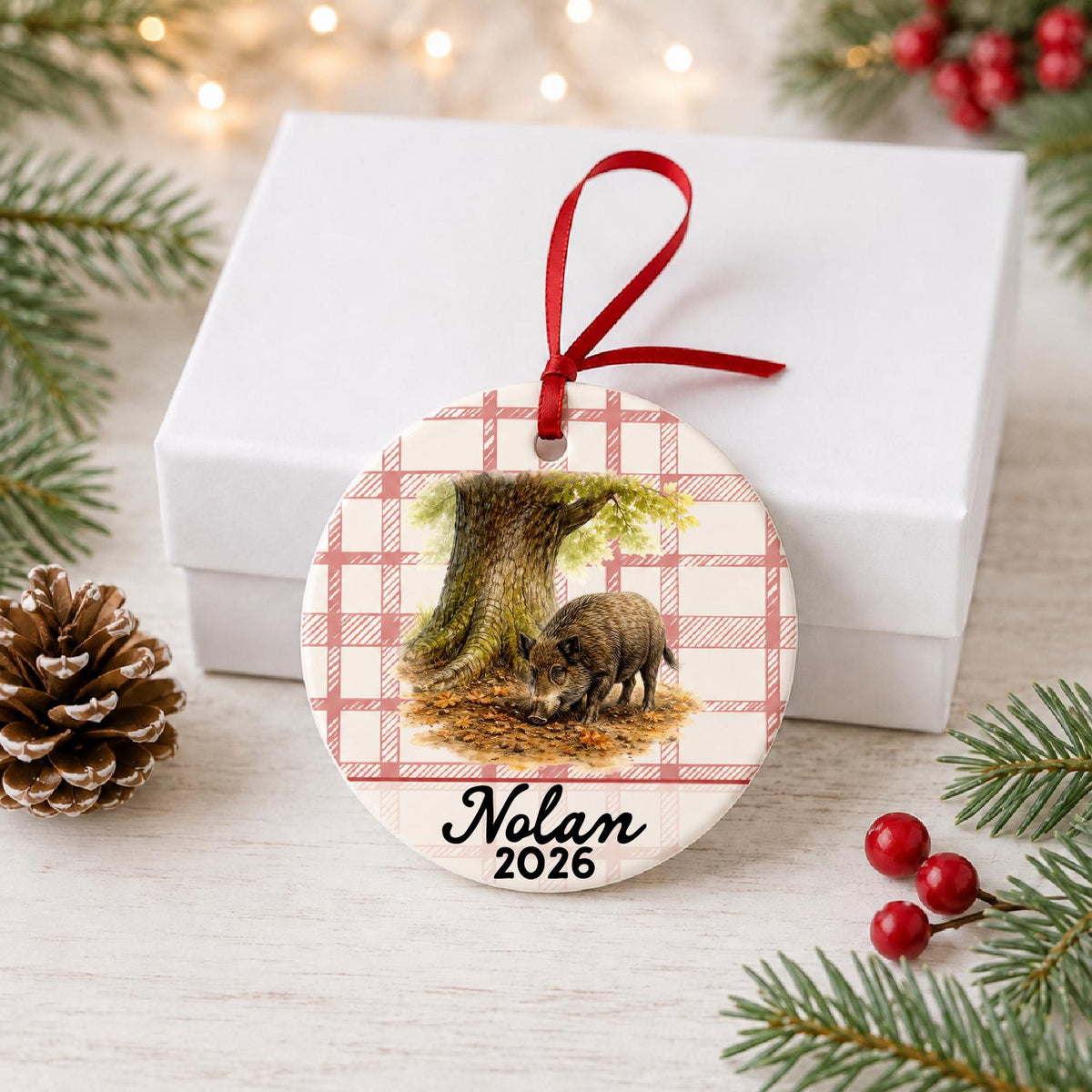 Personalized wild boar ceramic ornament with name and year on red plaid in front of a white gift box with pinecones and red berries