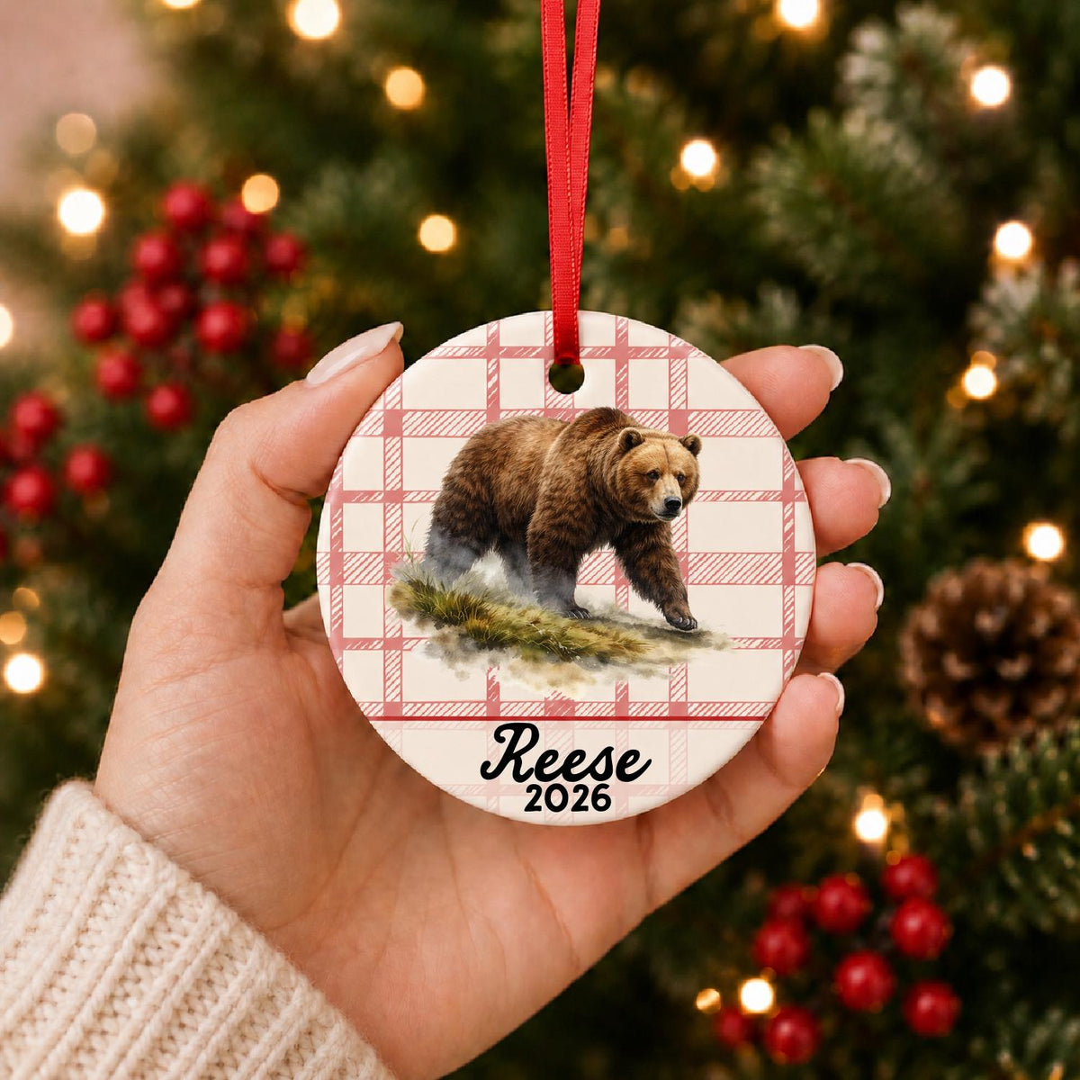 Hand holding the personalized brown bear ceramic ornament with name and year on red plaid in front of a Christmas tree