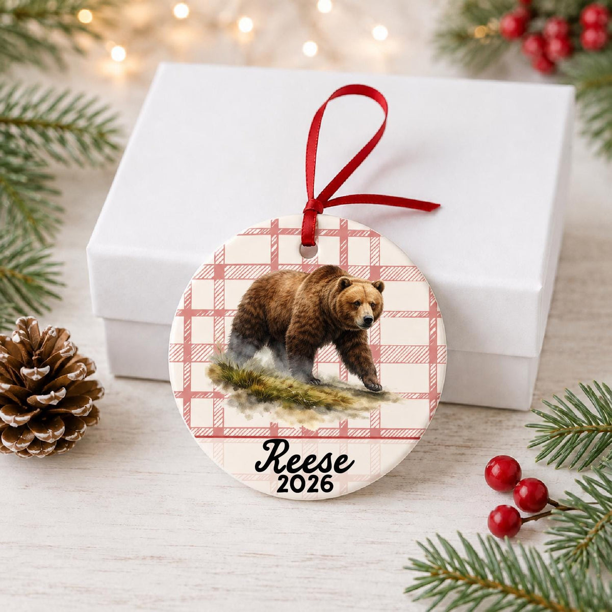 Personalized brown bear ceramic ornament with name and year on red plaid in front of a white gift box with pinecones and red berries