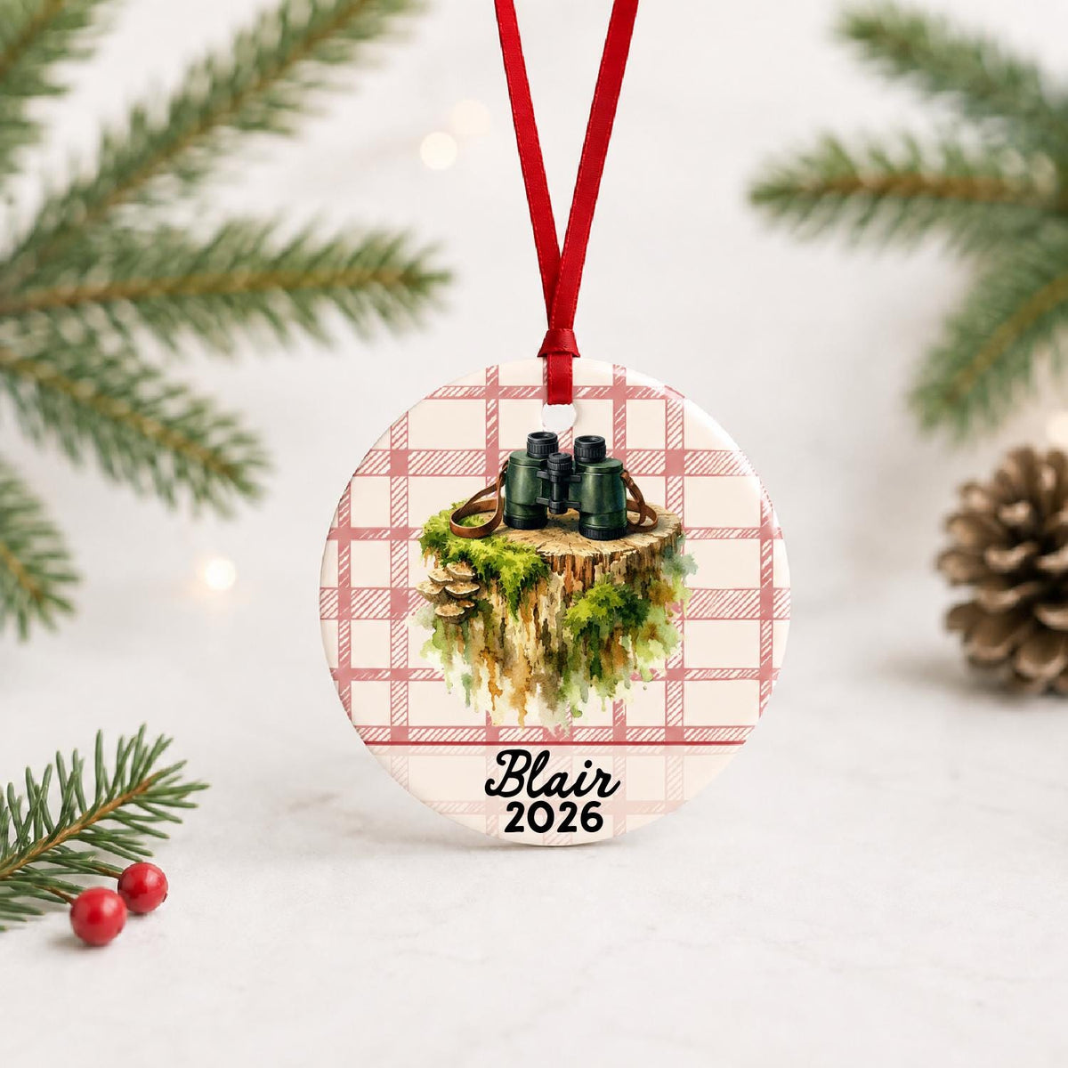 Personalized binoculars ceramic ornament with name and year on red plaid with red ribbon on a white surface with pine branches and pinecones