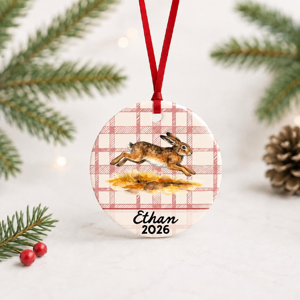 Personalized brown hare ceramic ornament with name and year on red plaid with red ribbon on a white surface with pine branches and pinecones