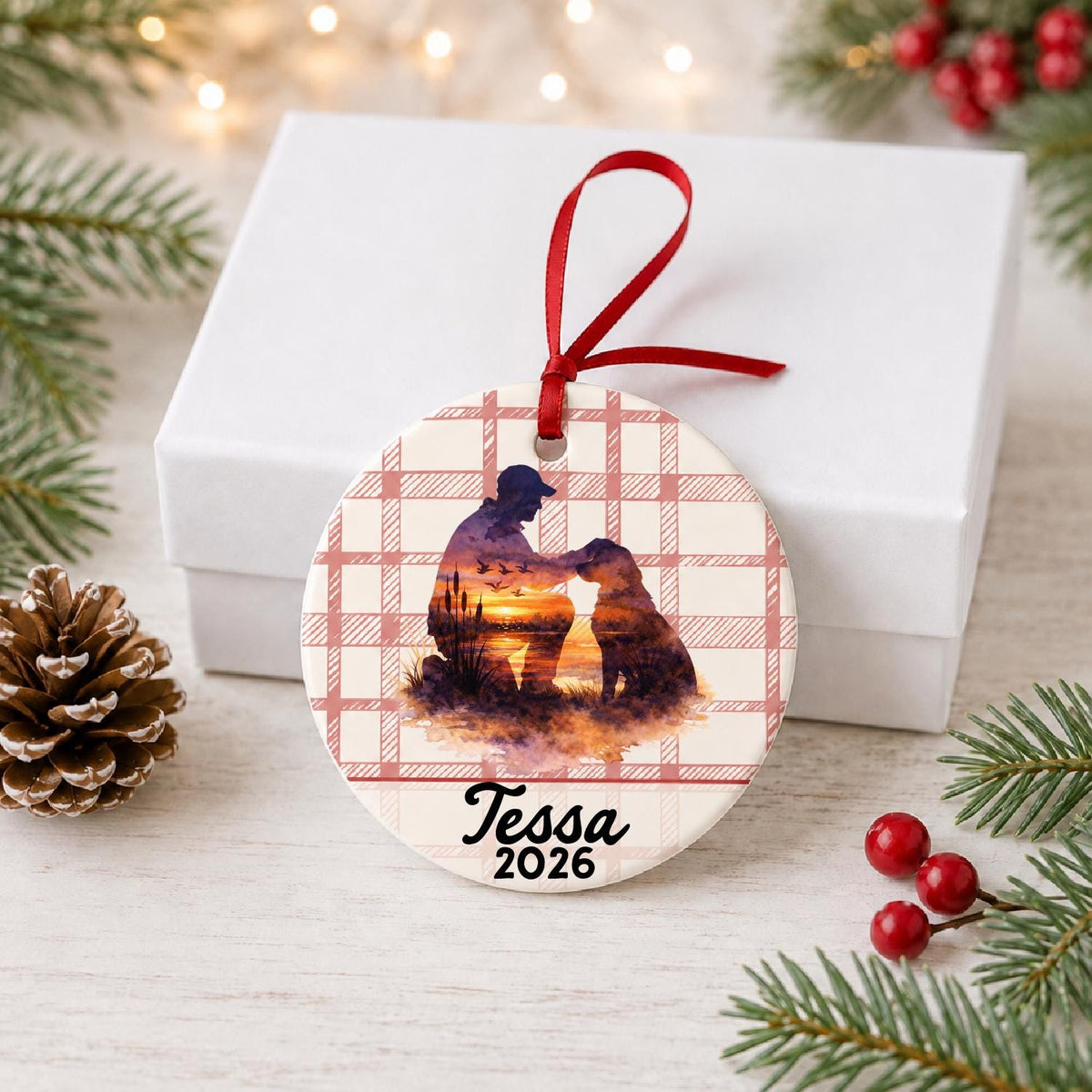 Personalized hunter and dog ceramic ornament with name and year on red plaid in front of a white gift box with pinecones and red berries