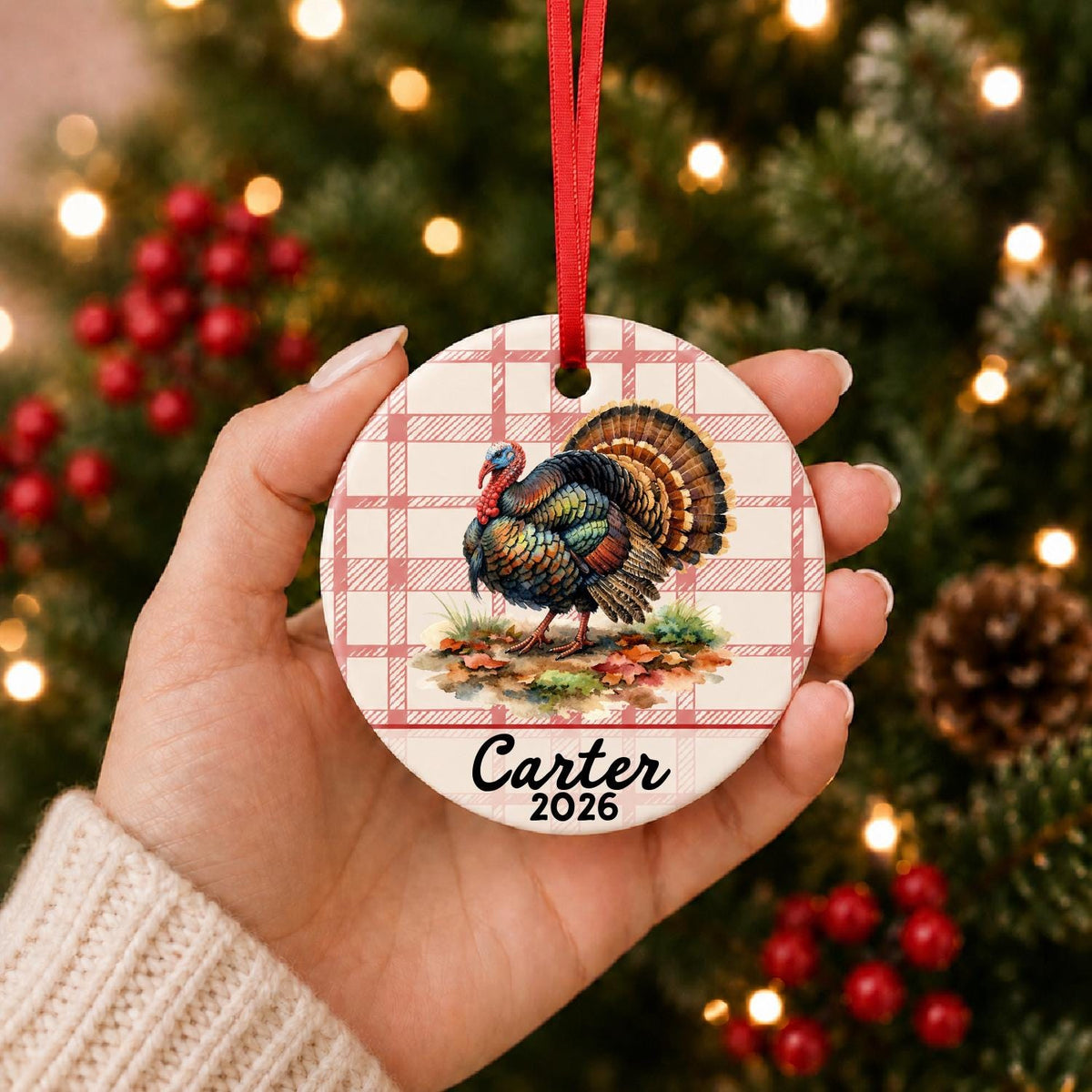 Hand holding the personalized wild turkey ceramic ornament with name and year on red plaid in front of a Christmas tree