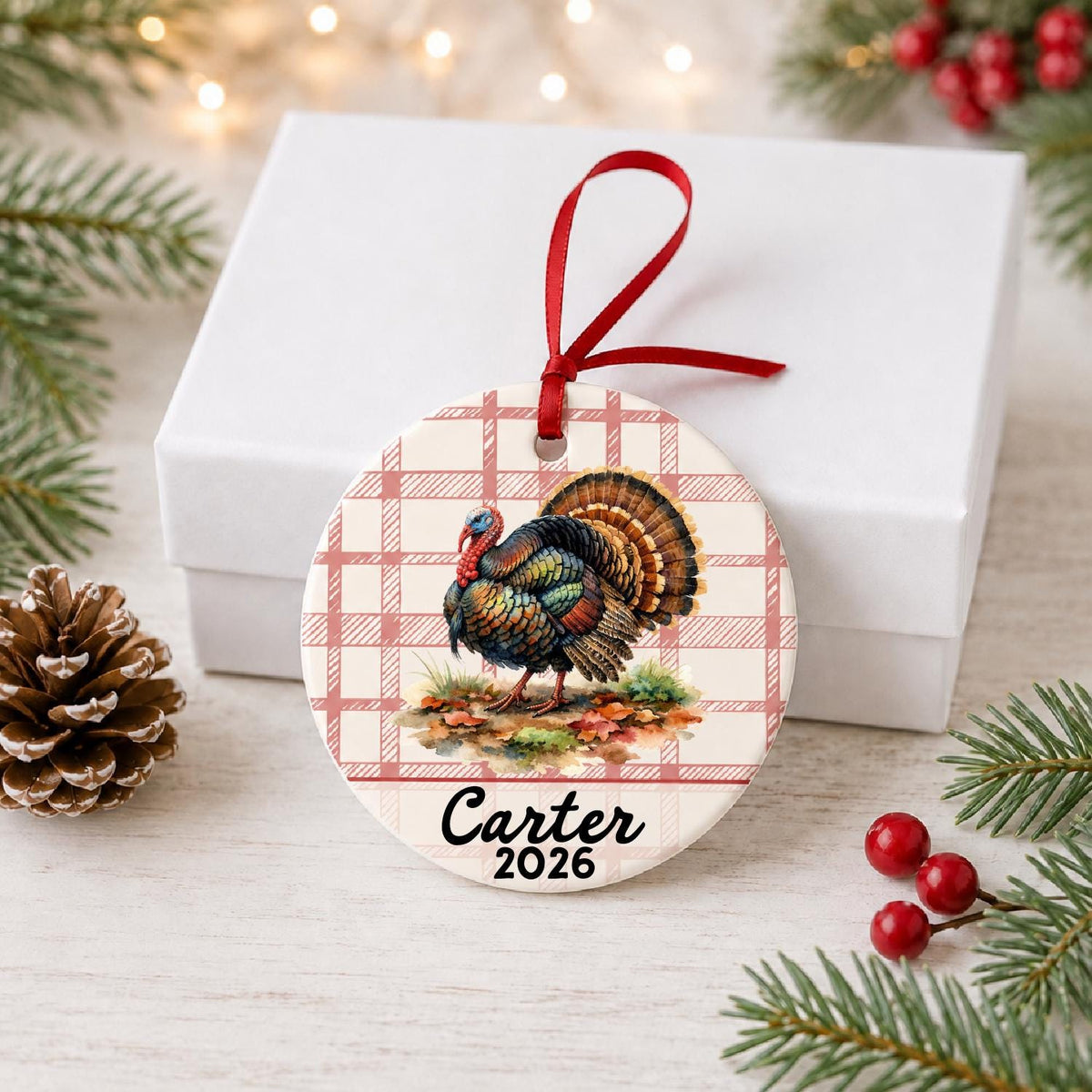 Personalized wild turkey ceramic ornament with name and year on red plaid in front of a white gift box with pinecones and red berries