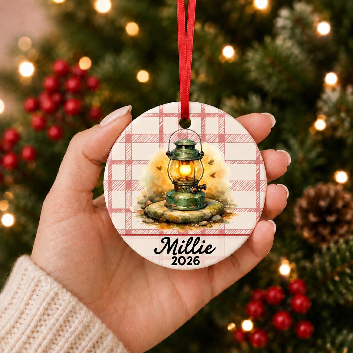 Hand holding the personalized camping lantern ceramic ornament with name and year on red plaid in front of a Christmas tree