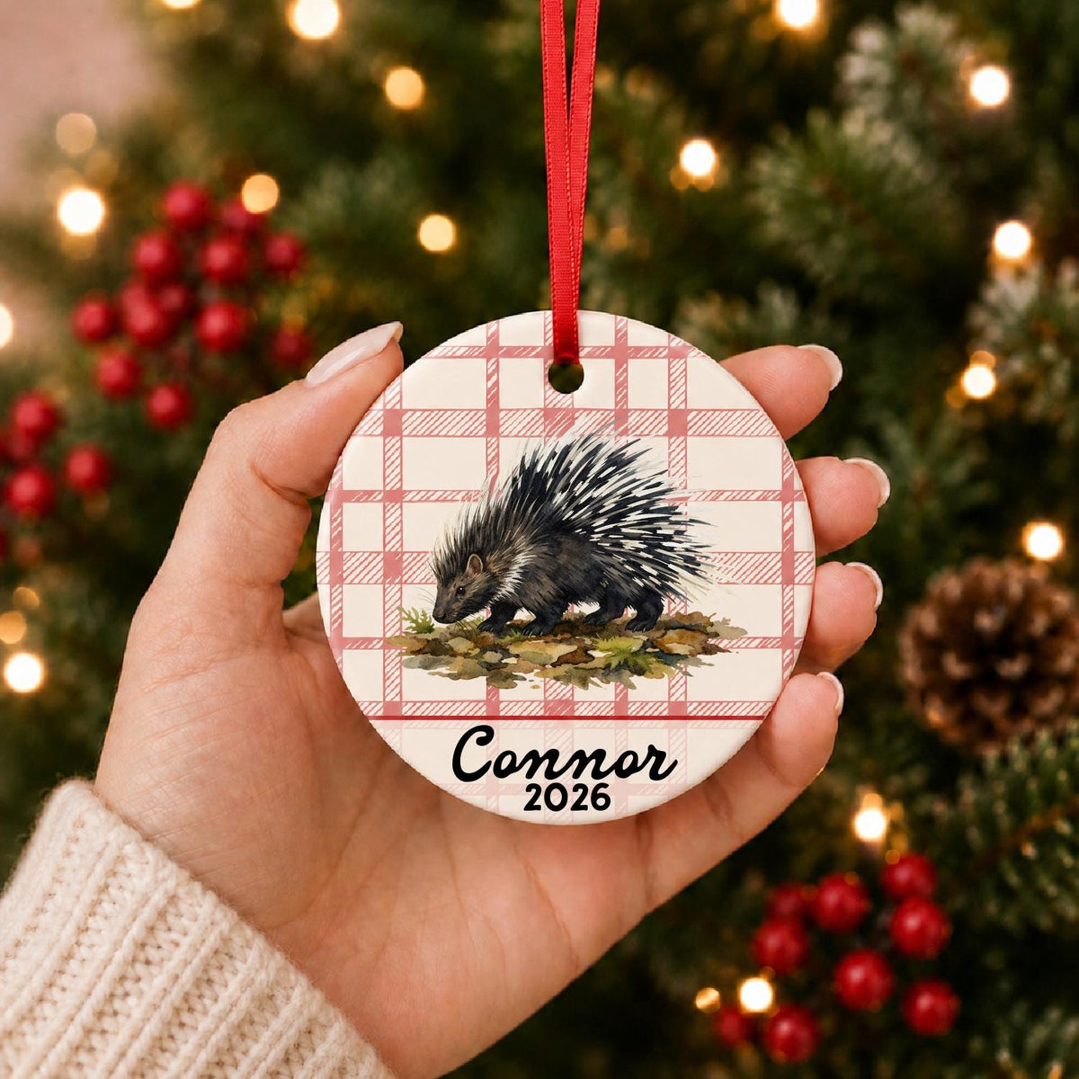 Hand holding the personalized porcupine ceramic ornament with name and year on red plaid in front of a Christmas tree
