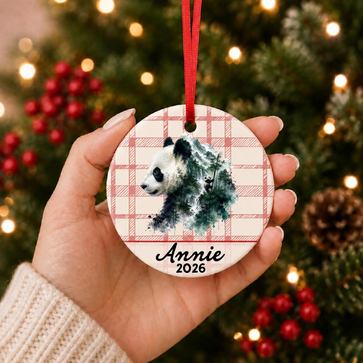 Hand holding the personalized giant panda ceramic ornament with name and year on red plaid in front of a Christmas tree