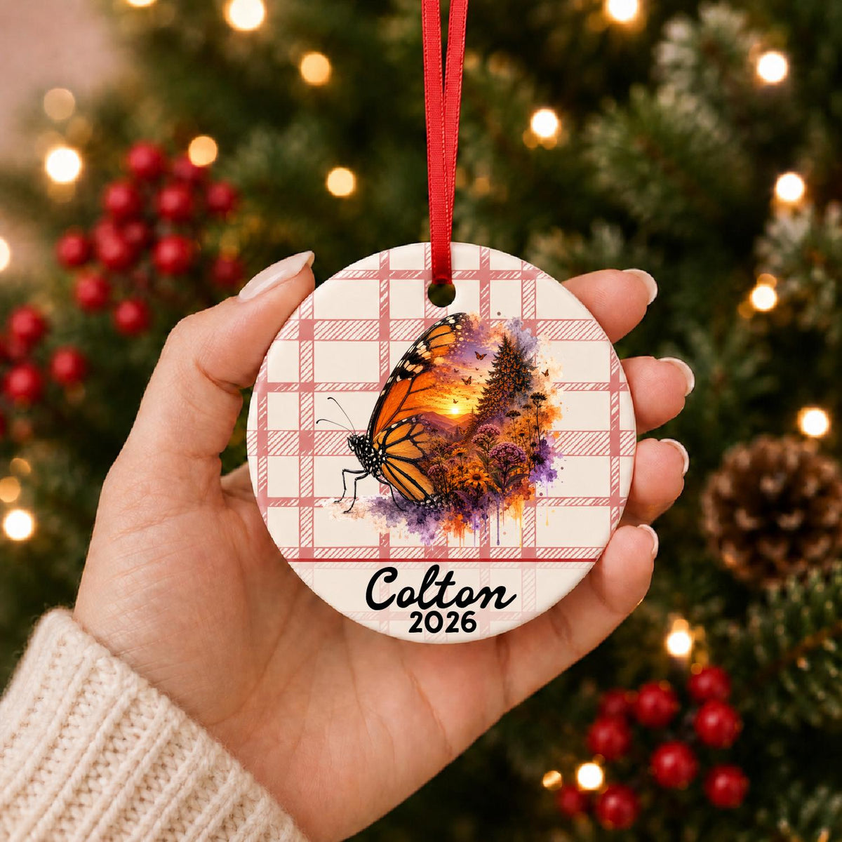 Hand holding the personalized monarch butterfly ceramic ornament with name and year on red plaid in front of a Christmas tree