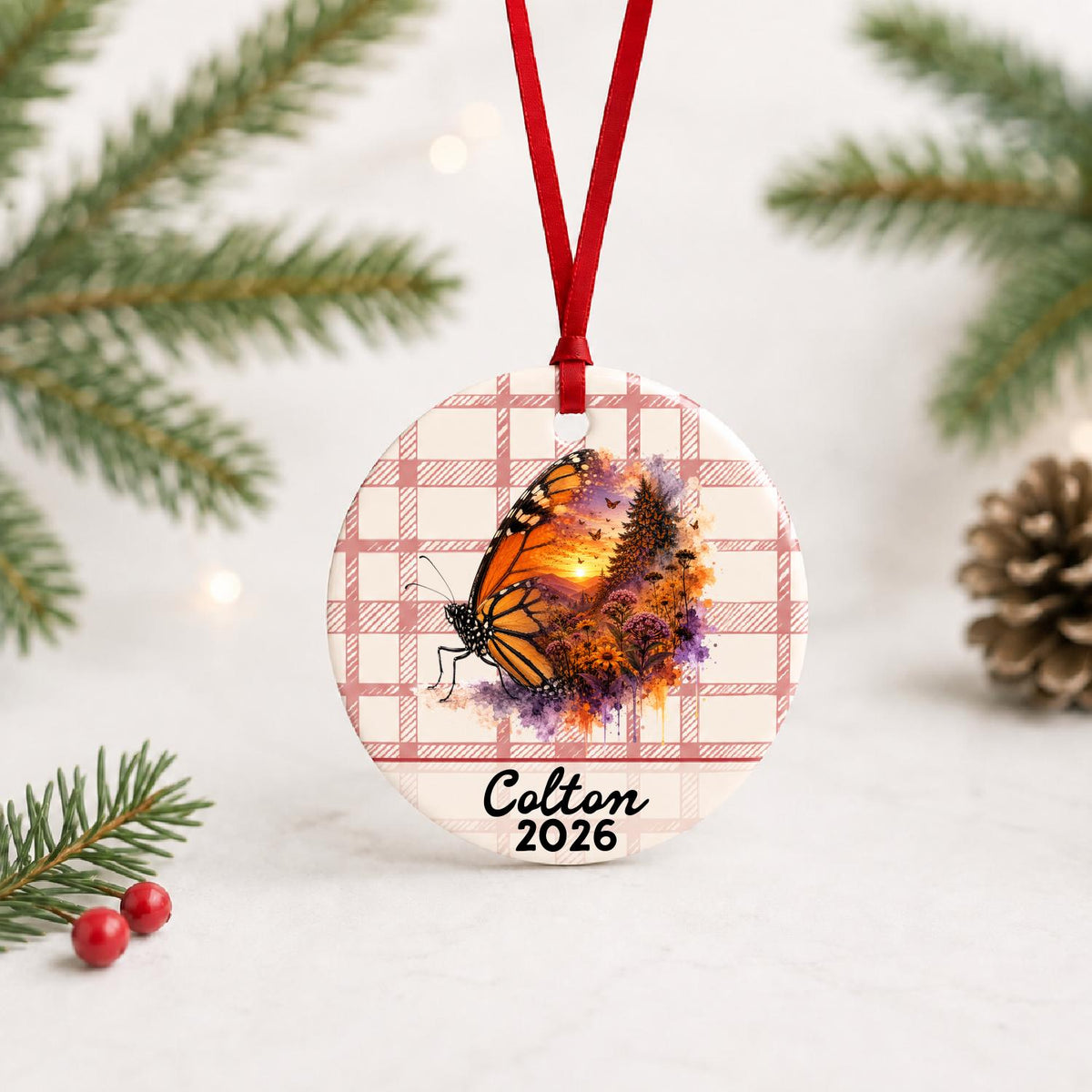 Personalized monarch butterfly ceramic ornament with name and year on red plaid with red ribbon on a white surface with pine branches and pinecones