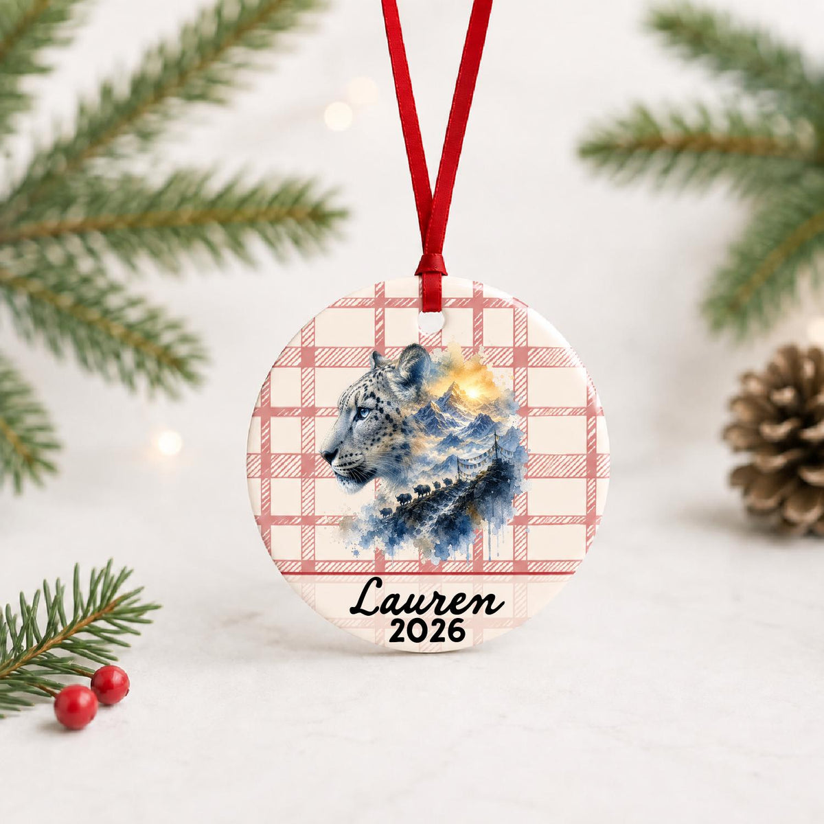 Personalized snow leopard ceramic ornament with name and year on red plaid with red ribbon on a white surface with pine branches and pinecones