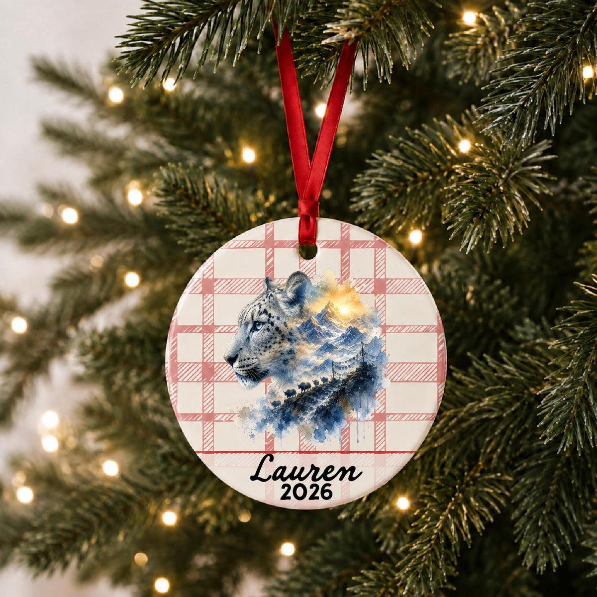 Personalized snow leopard ceramic ornament with name and year on red plaid hanging on a lit Christmas tree
