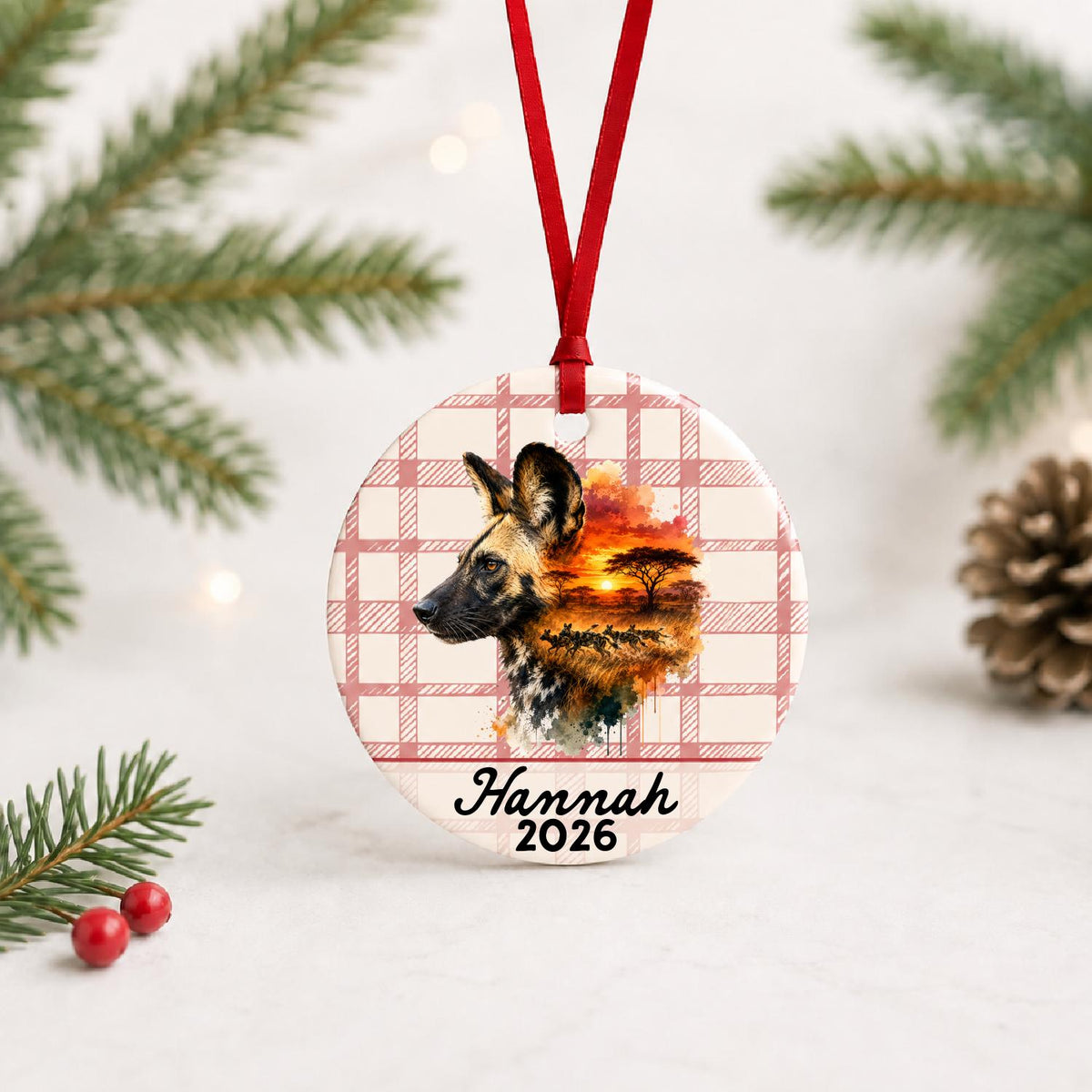 Personalized african wild dog ceramic ornament with name and year on red plaid with red ribbon on a white surface with pine branches and pinecones