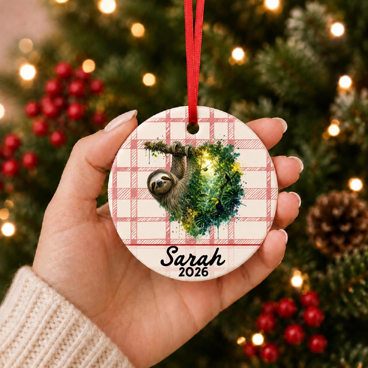 Hand holding the personalized sloth ceramic ornament with name and year on red plaid in front of a Christmas tree
