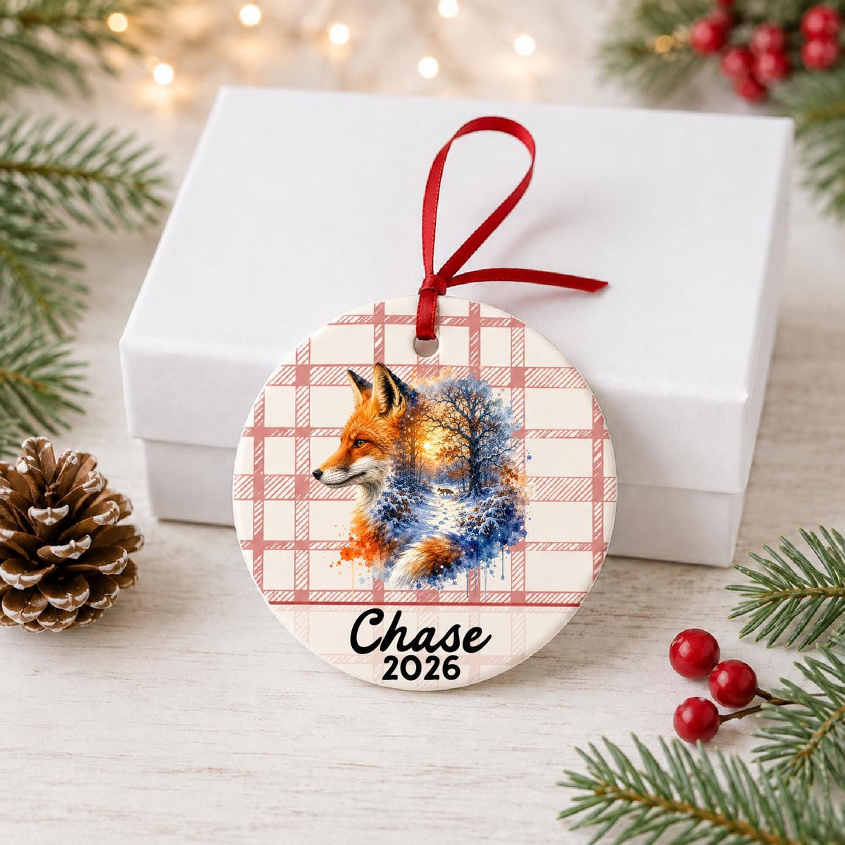 Personalized red fox ceramic ornament with name and year on red plaid in front of a white gift box with pinecones and red berries