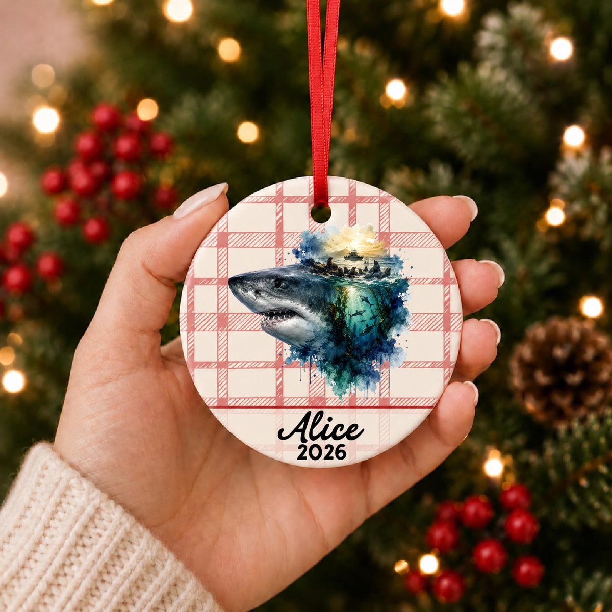 Hand holding the personalized great white shark ceramic ornament with name and year on red plaid in front of a Christmas tree
