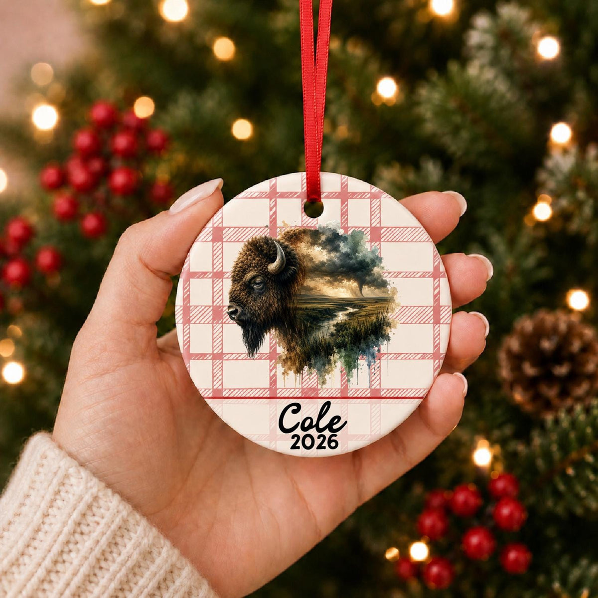 Hand holding the personalized american bison ceramic ornament with name and year on red plaid in front of a Christmas tree