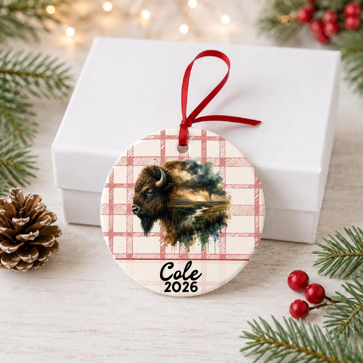 Personalized american bison ceramic ornament with name and year on red plaid in front of a white gift box with pinecones and red berries
