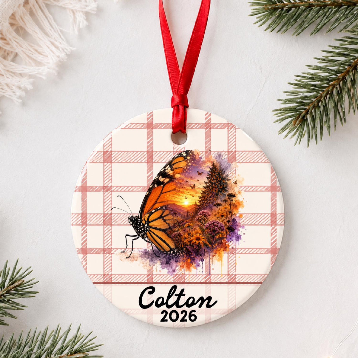 Personalized monarch butterfly ceramic ornament with name and year on red plaid styled with pine branches on a soft white background