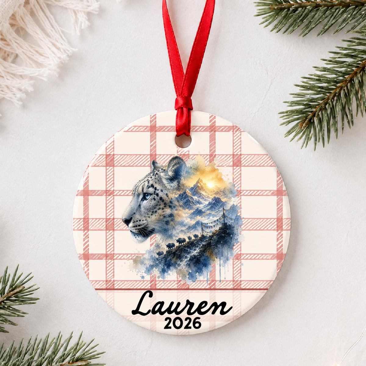 Personalized snow leopard ceramic ornament with name and year on red plaid styled with pine branches on a soft white background