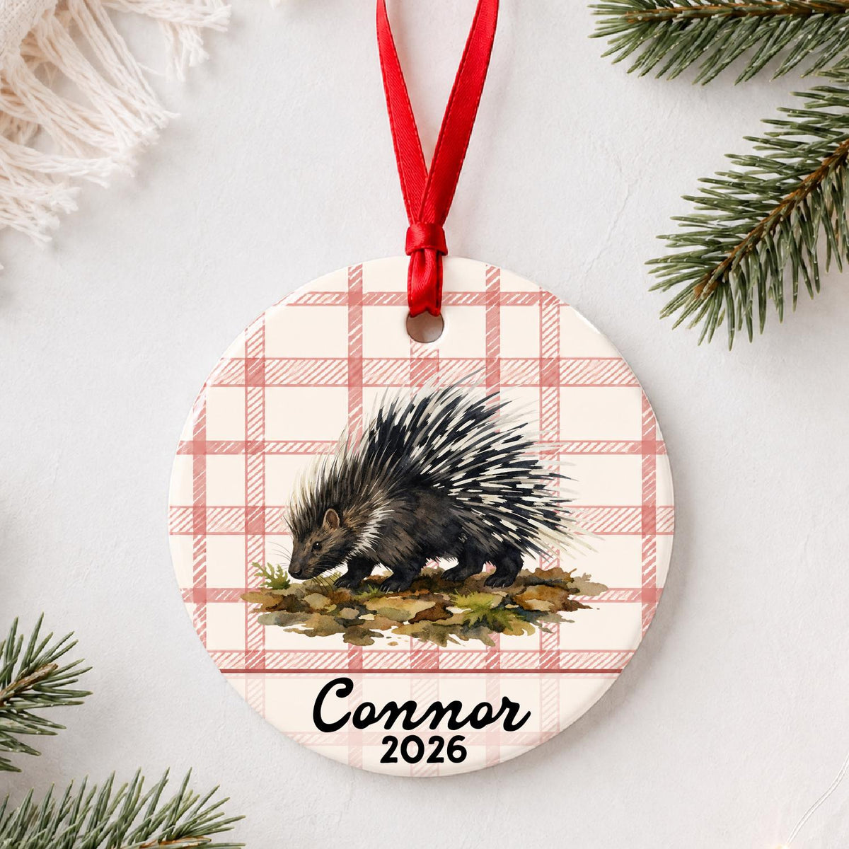 Personalized porcupine ceramic ornament with name and year on red plaid styled with pine branches on a soft white background