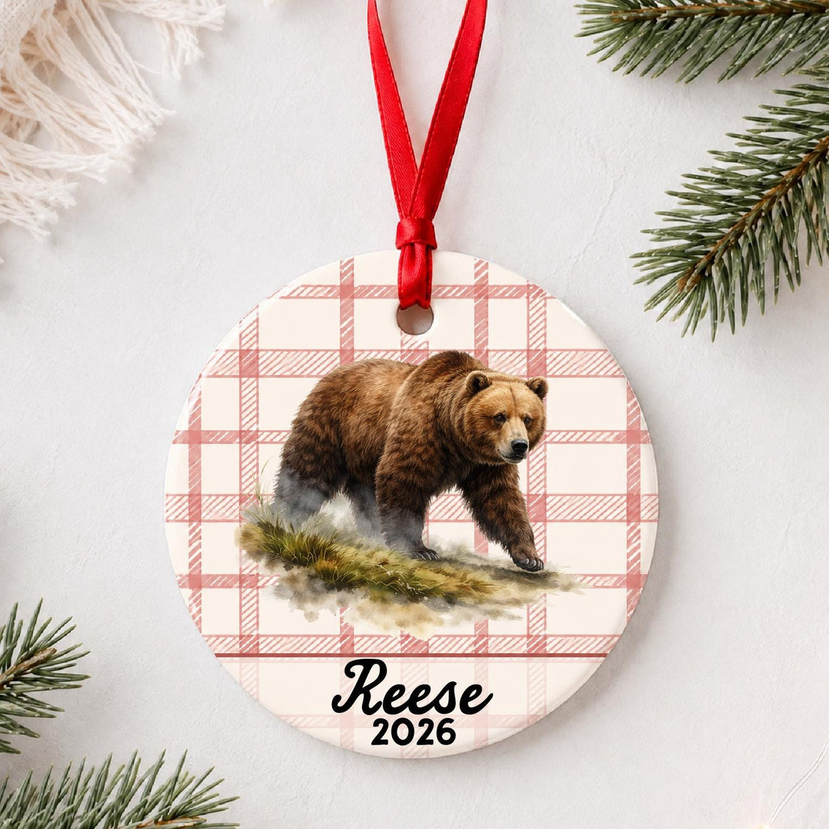 Personalized brown bear ceramic ornament with name and year on red plaid styled with pine branches on a soft white background