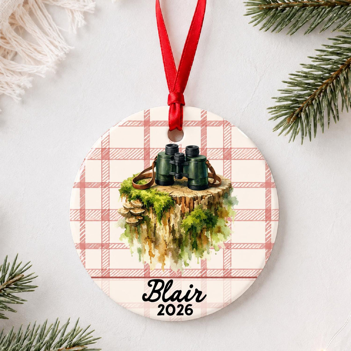 Personalized binoculars ceramic ornament with name and year on red plaid styled with pine branches on a soft white background