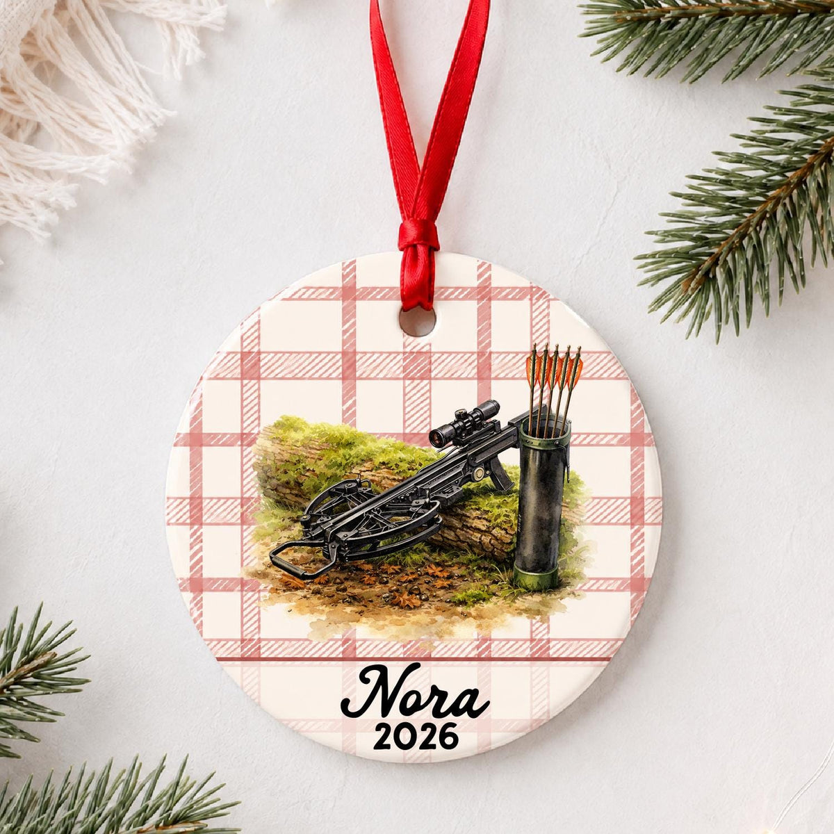 Personalized crossbow ceramic ornament with name and year on red plaid styled with pine branches on a soft white background