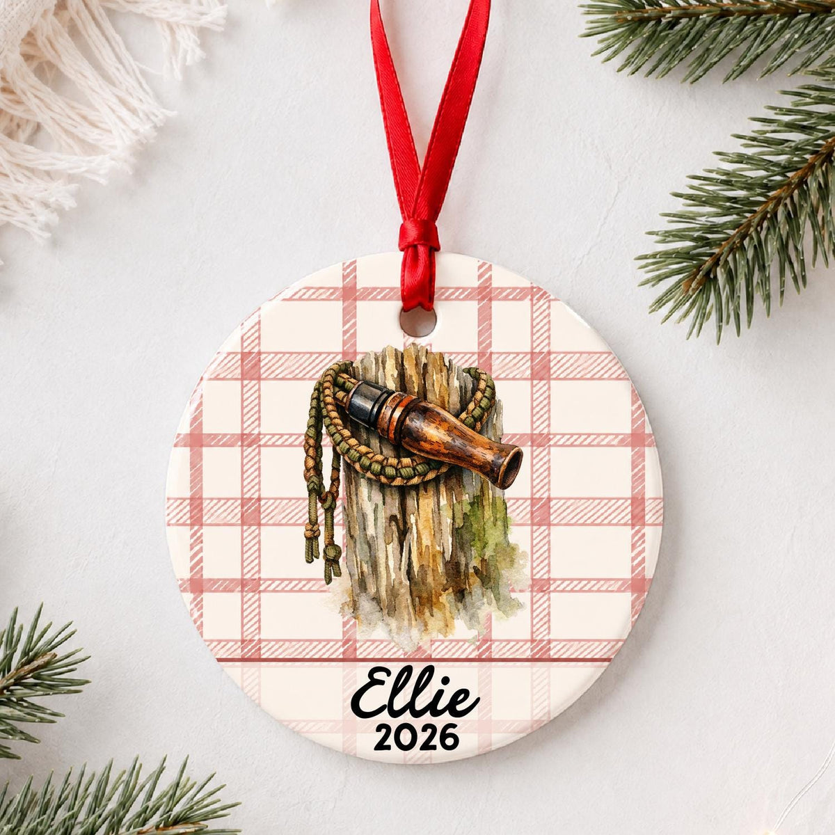 Personalized duck call ceramic ornament with name and year on red plaid styled with pine branches on a soft white background