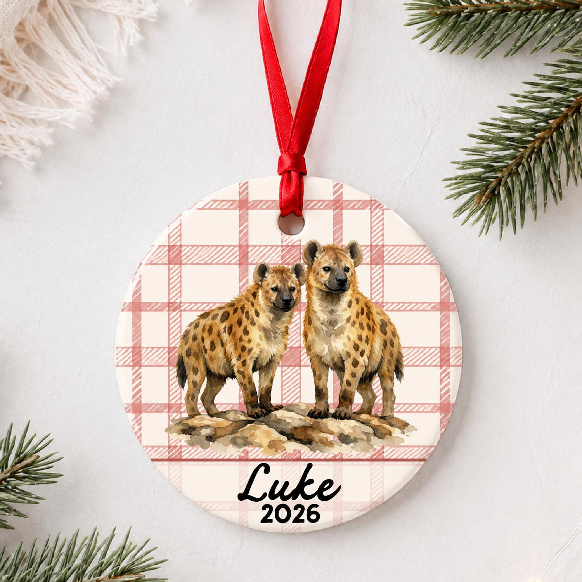 Personalized spotted hyena ceramic ornament with name and year on red plaid styled with pine branches on a soft white background