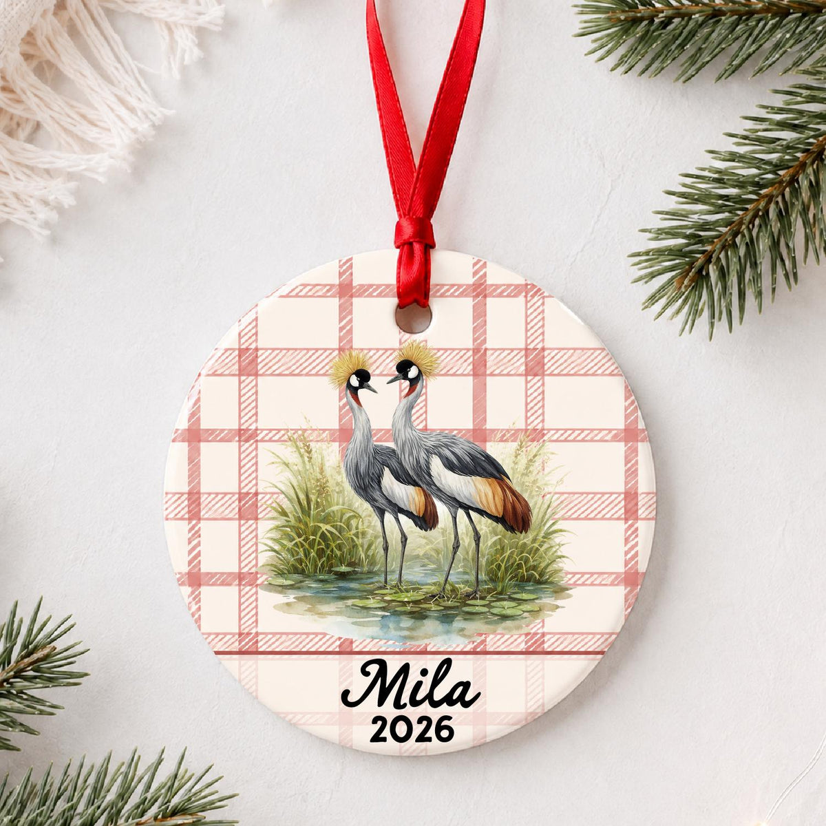 Personalized grey crowned crane ceramic ornament with name and year on red plaid styled with pine branches on a soft white background