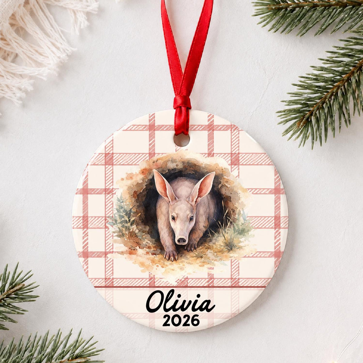Personalized aardvark ceramic ornament with name and year on red plaid styled with pine branches on a soft white background