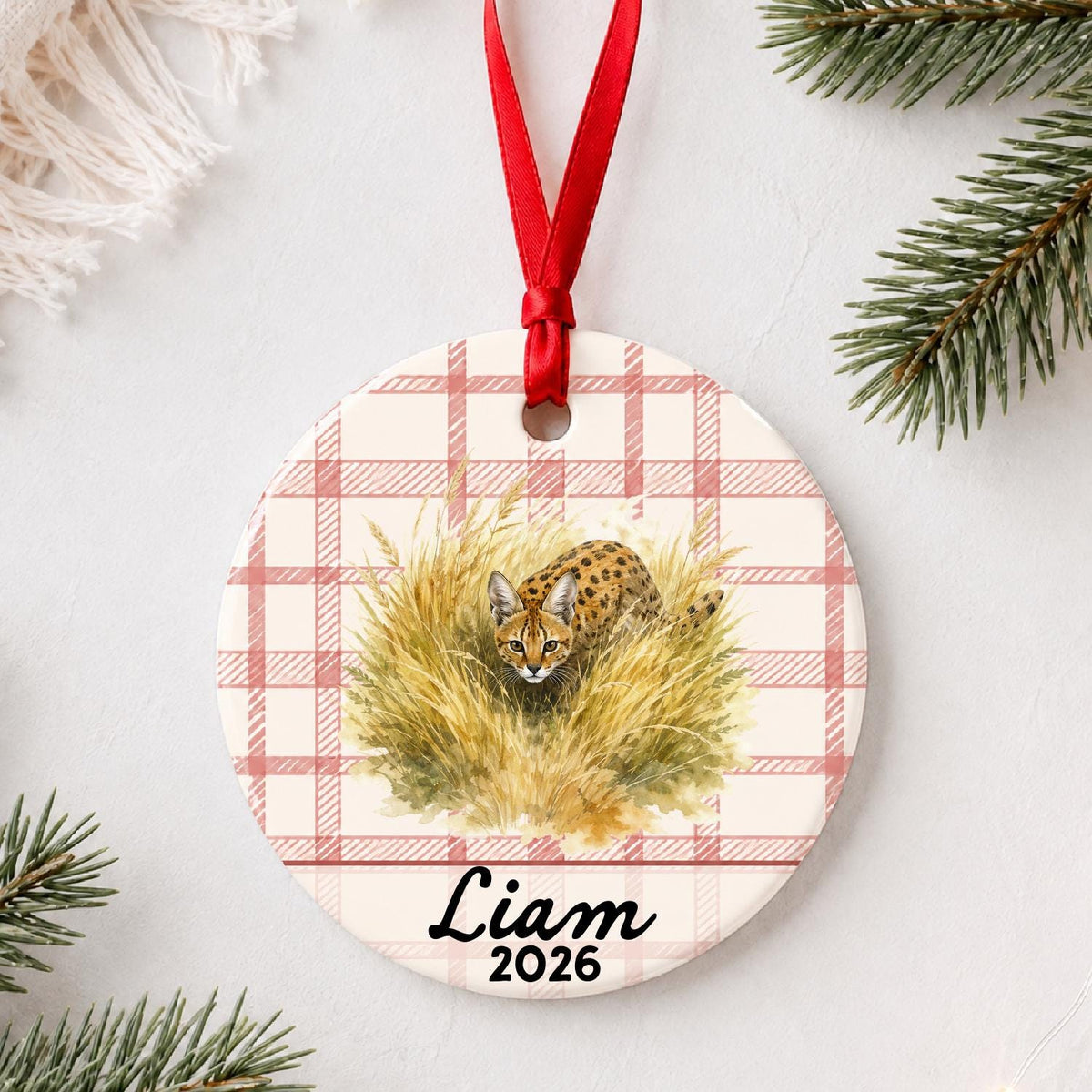 Personalized serval ceramic ornament with name and year on red plaid styled with pine branches on a soft white background