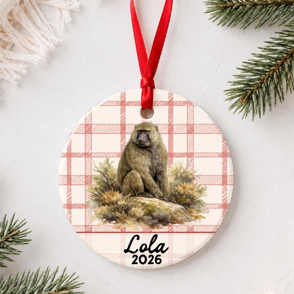 Personalized baboon ceramic ornament with name and year on red plaid styled with pine branches on a soft white background