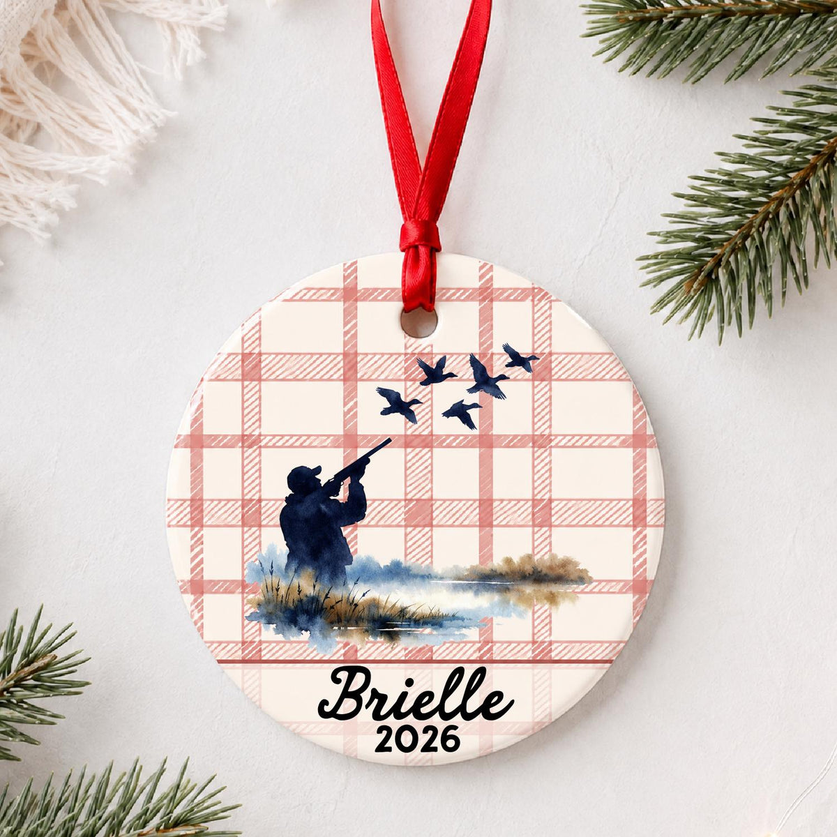 Personalized duck hunting ceramic ornament with name and year on red plaid styled with pine branches on a soft white background