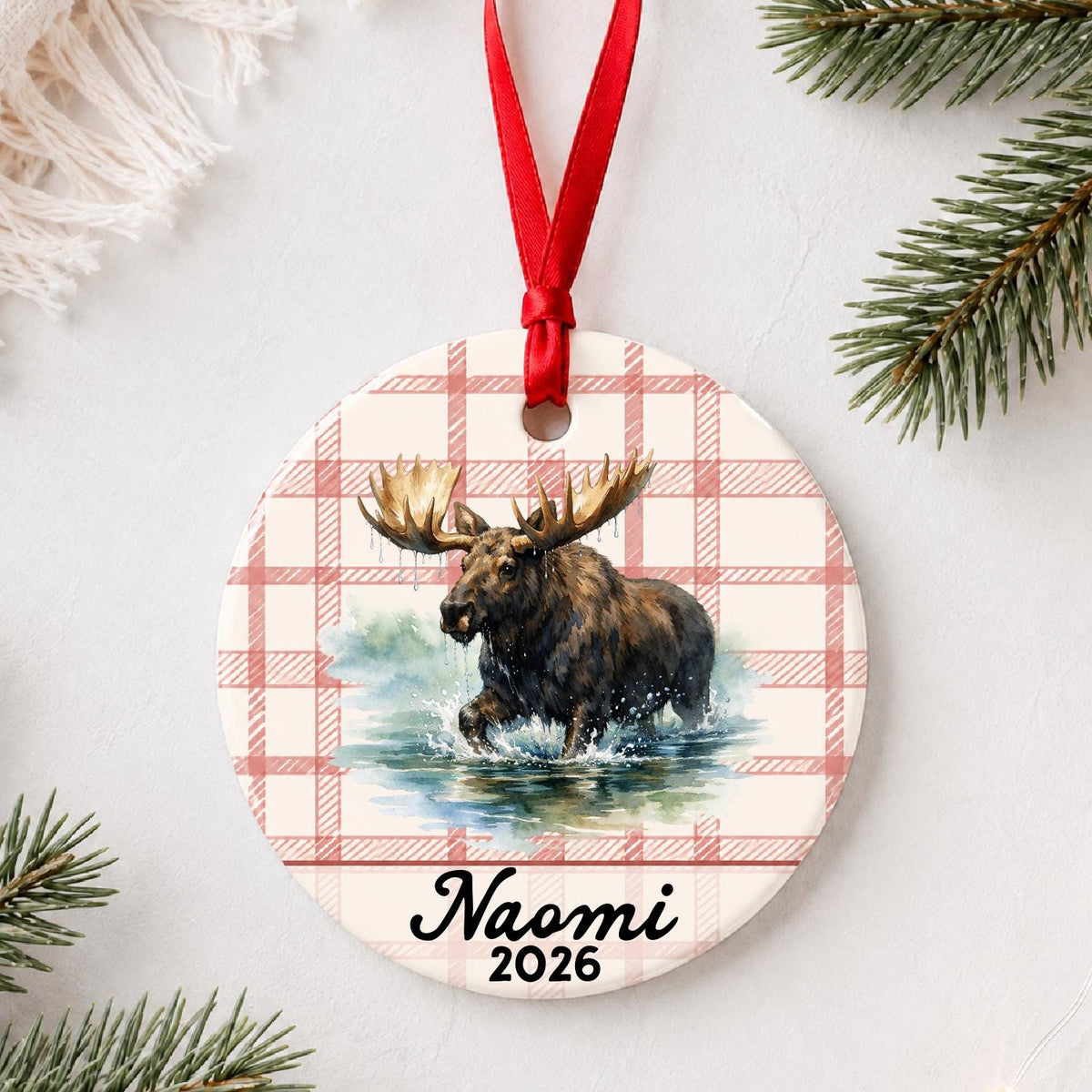 Personalized bull moose ceramic ornament with name and year on red plaid styled with pine branches on a soft white background