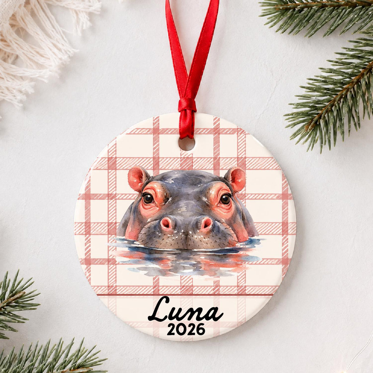 Personalized hippopotamus ceramic ornament with name and year on red plaid styled with pine branches on a soft white background