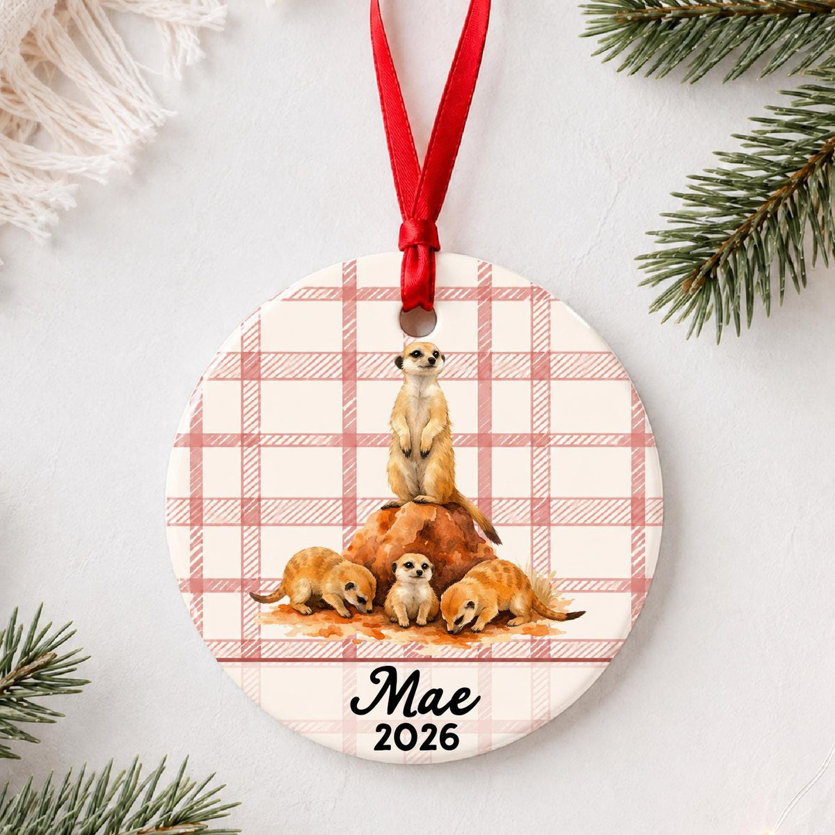 Personalized meerkat ceramic ornament with name and year on red plaid styled with pine branches on a soft white background