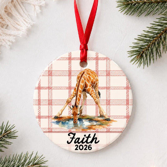 Personalized giraffe ceramic ornament with name and year on red plaid styled with pine branches on a soft white background