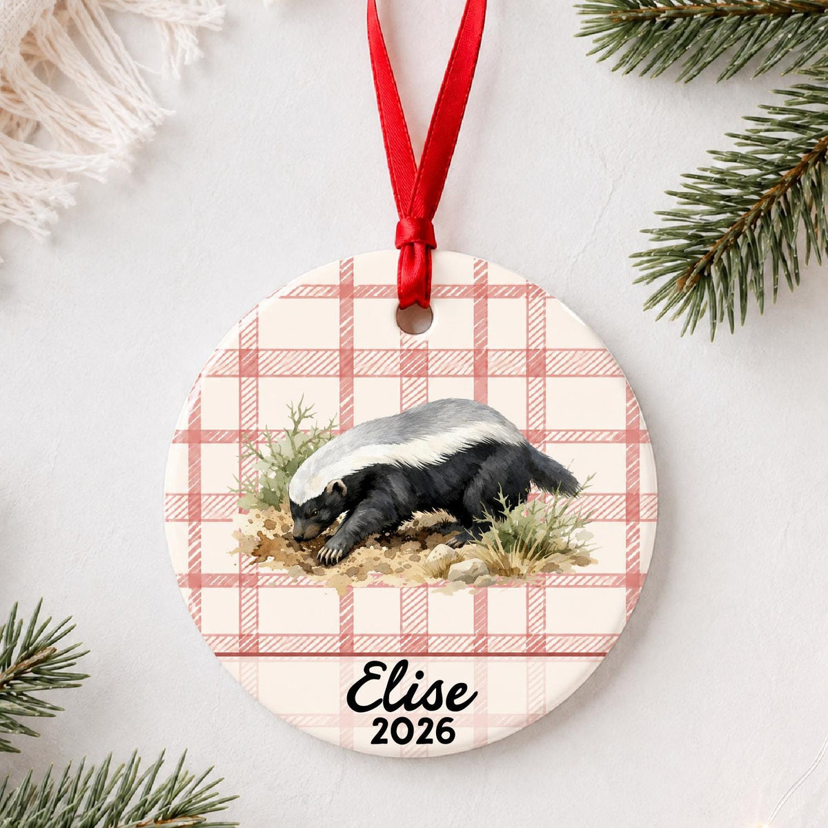 Personalized honey badger ceramic ornament with name and year on red plaid styled with pine branches on a soft white background