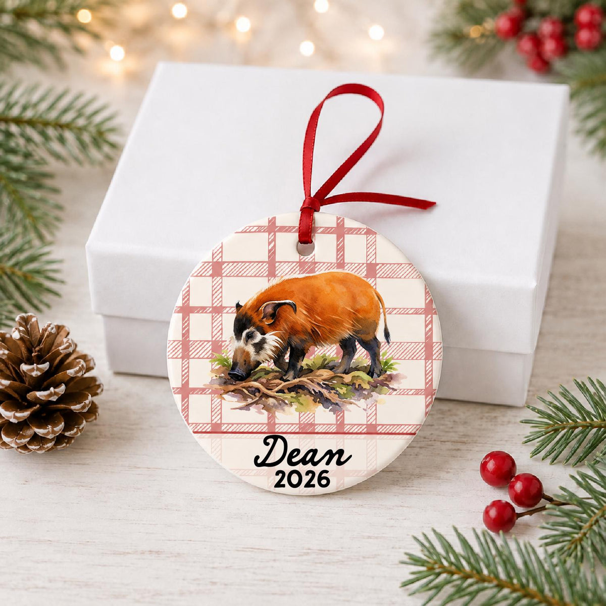 Personalized red river hog ceramic ornament with name and year on red plaid in front of a white gift box with pinecones and red berries