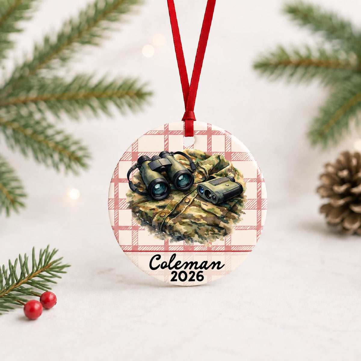 Personalized binoculars and rangefinder ceramic ornament with name and year on red plaid with red ribbon on a white surface with pine branches and pinecones