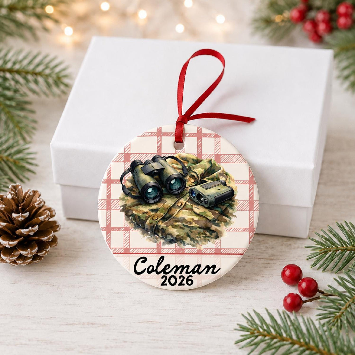 Personalized binoculars and rangefinder ceramic ornament with name and year on red plaid in front of a white gift box with pinecones and red berries
