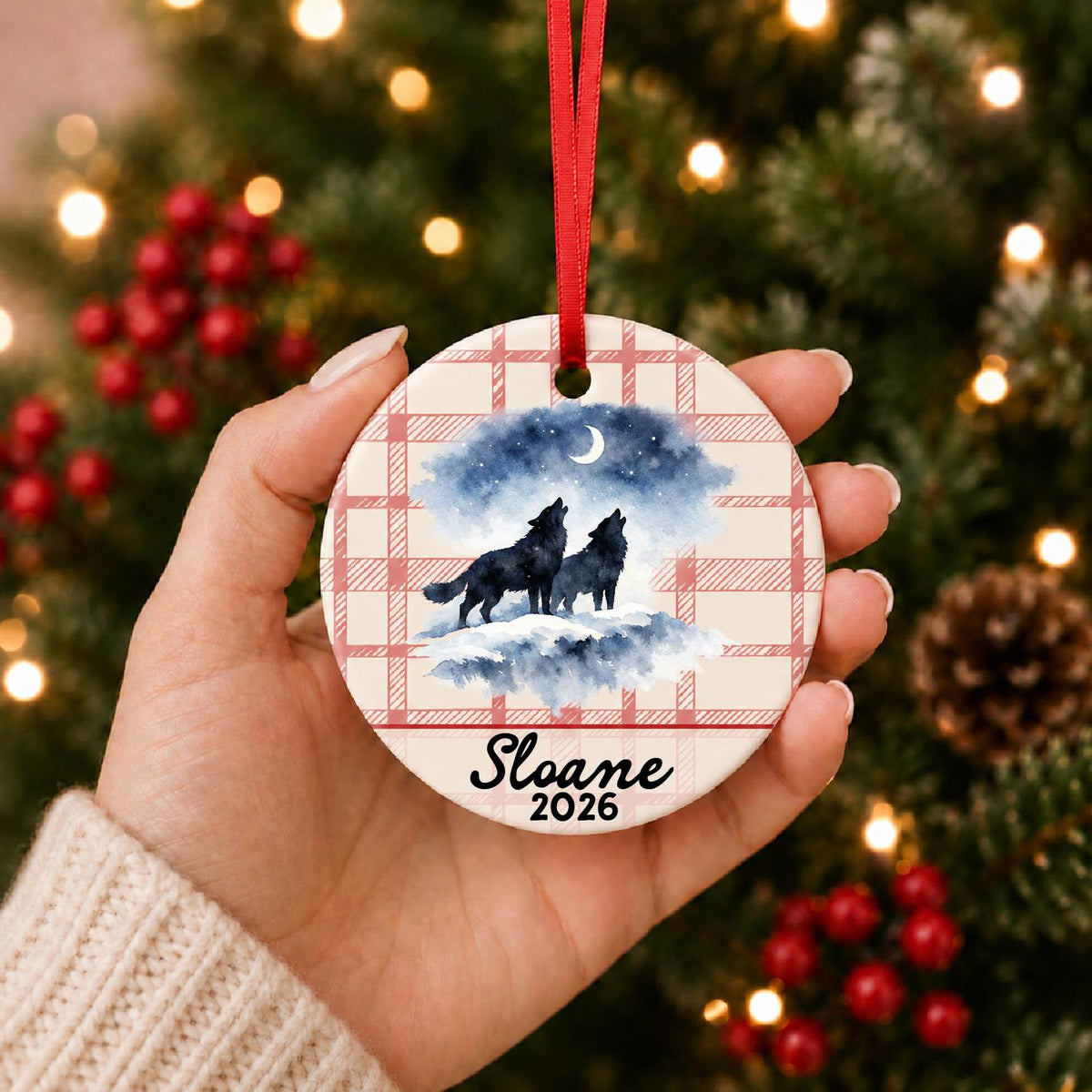 Hand holding the personalized howling wolf pair ceramic ornament with name and year on red plaid in front of a Christmas tree