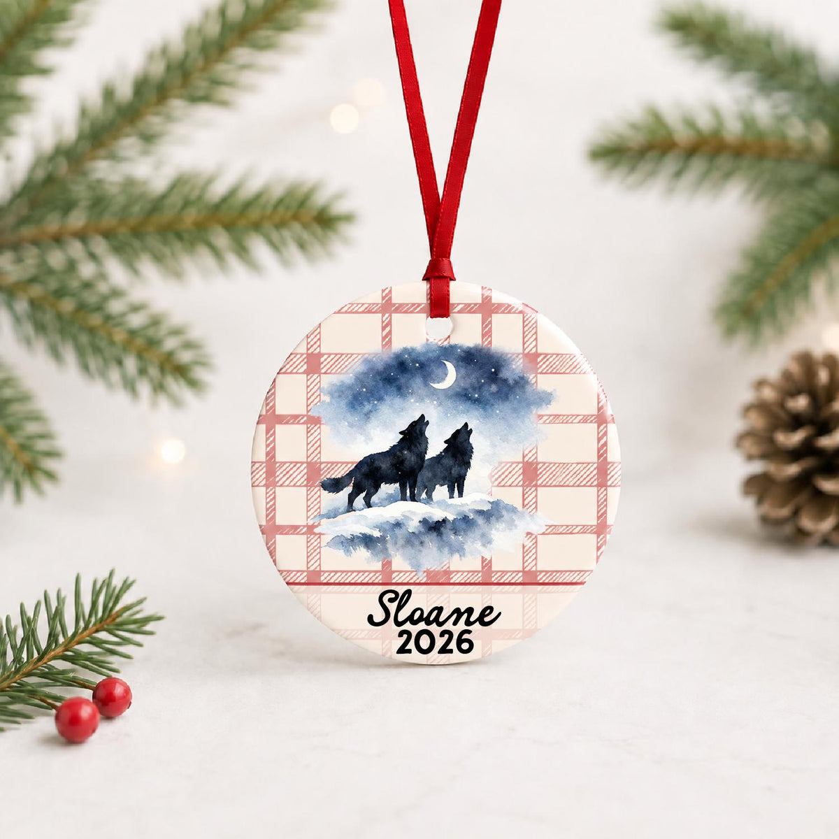 Personalized howling wolf pair ceramic ornament with name and year on red plaid with red ribbon on a white surface with pine branches and pinecones