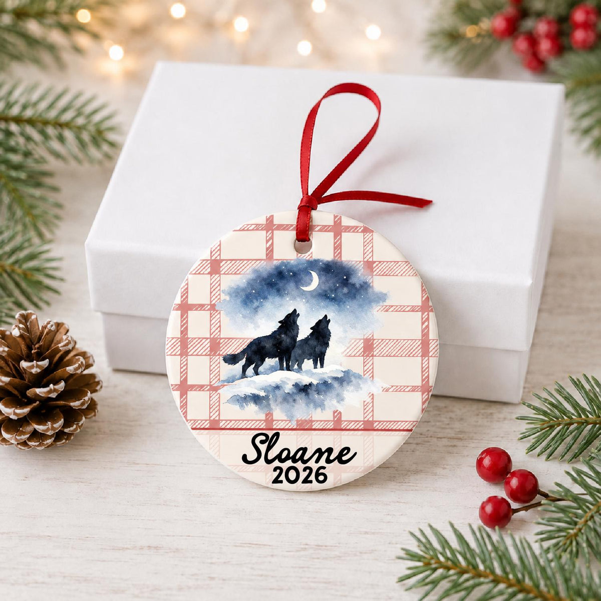 Personalized howling wolf pair ceramic ornament with name and year on red plaid in front of a white gift box with pinecones and red berries