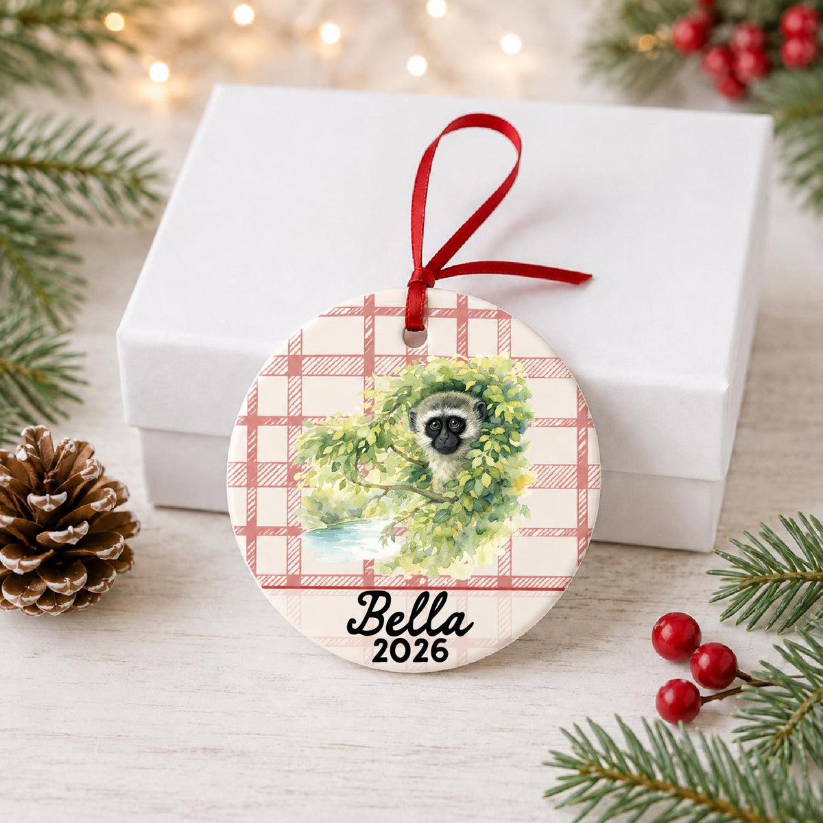 Personalized vervet monkey ceramic ornament with name and year on red plaid in front of a white gift box with pinecones and red berries