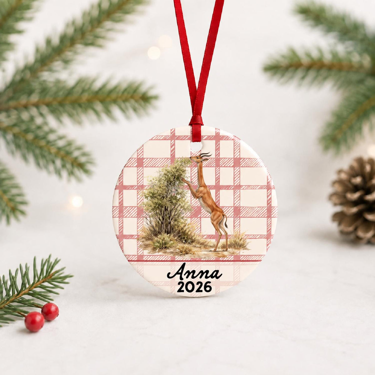 Personalized gerenuk ceramic ornament with name and year on red plaid with red ribbon on a white surface with pine branches and pinecones