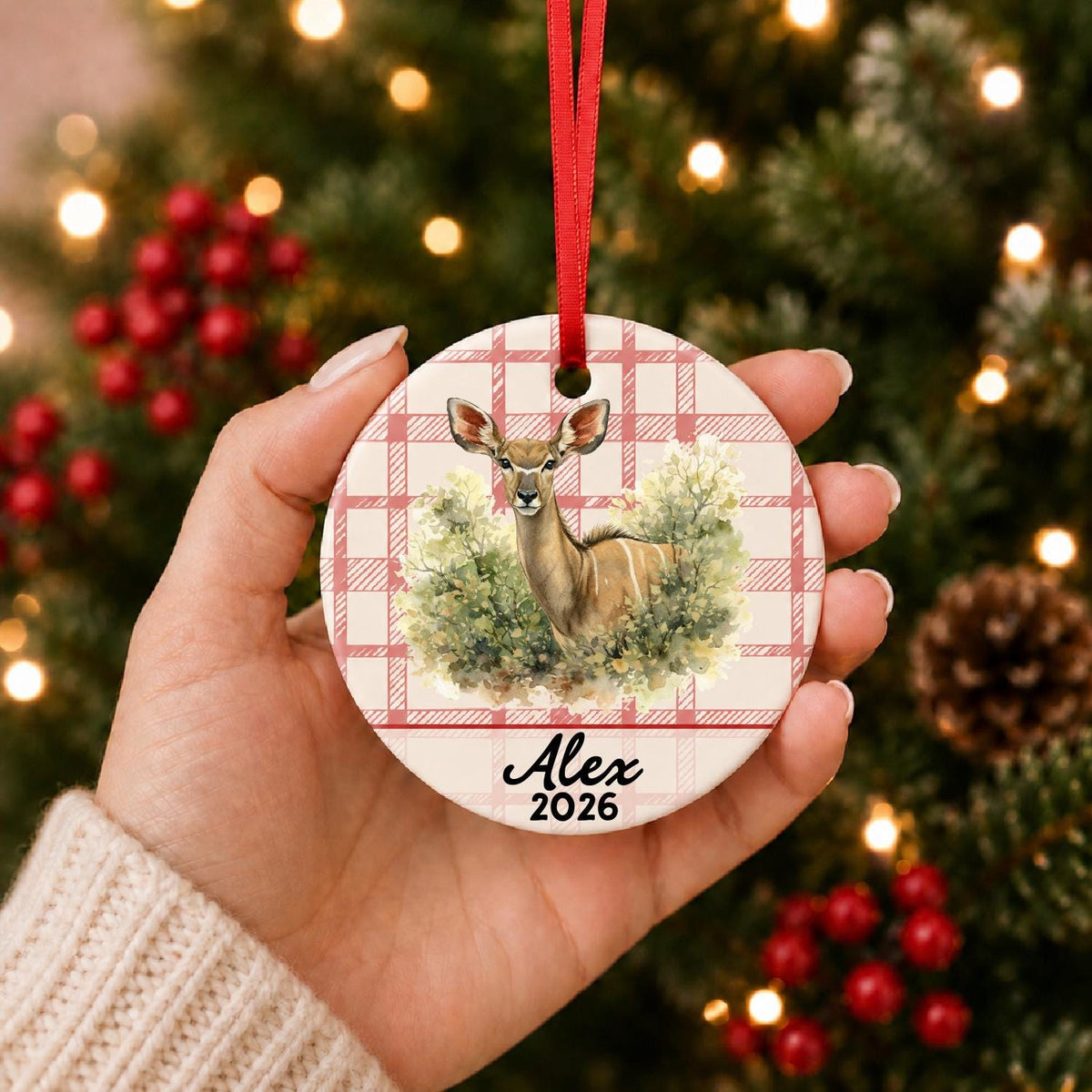 Hand holding the personalized kudu in woodland ceramic ornament with name and year on red plaid in front of a Christmas tree