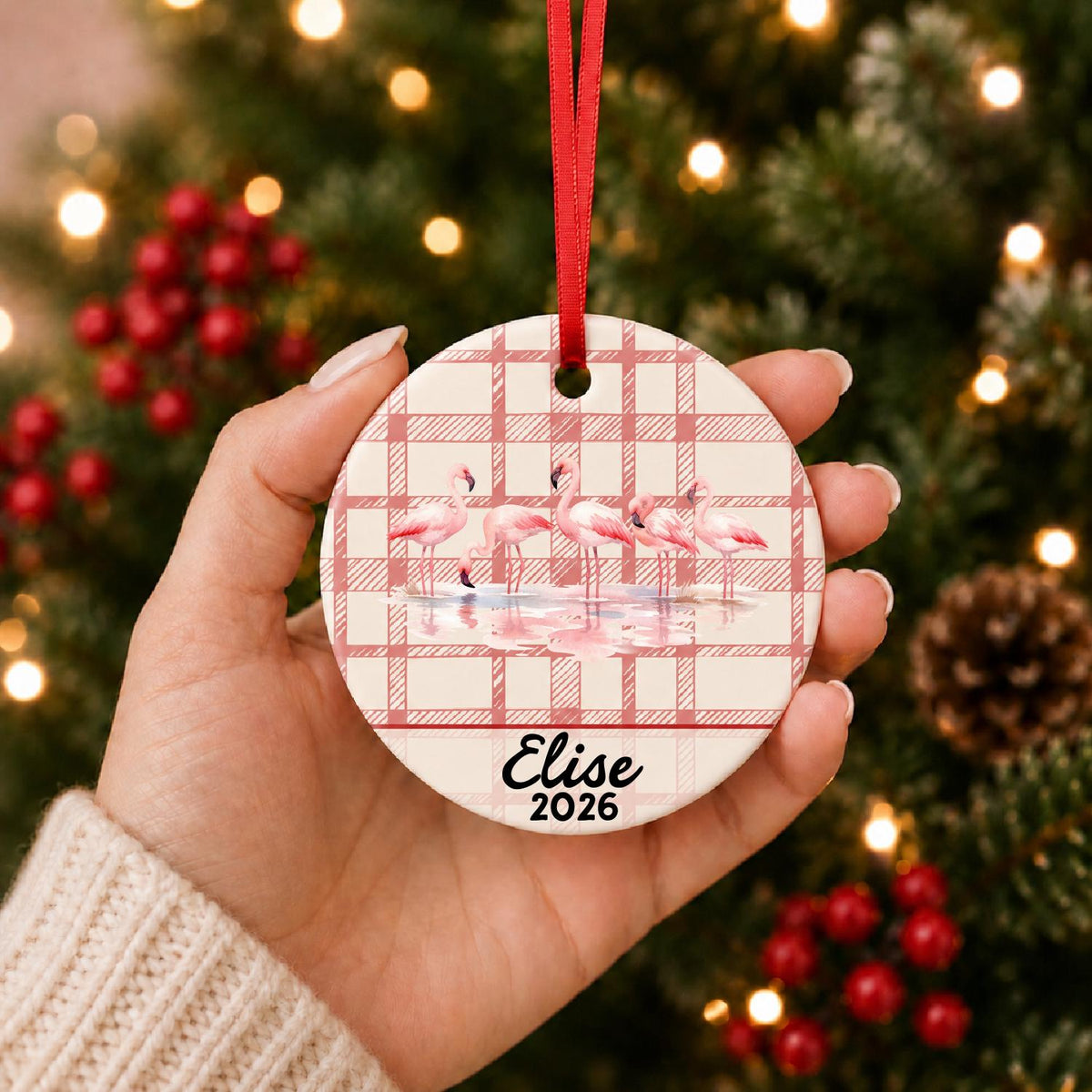 Hand holding the personalized flamingo flock ceramic ornament with name and year on red plaid in front of a Christmas tree